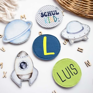 Könnte beinhalten: Eine Sammlung von Filz-Patches zum Thema Weltraum, darunter ein Planet, eine Rakete und ein UFO. Ein Patch hat den Buchstaben "L", ein anderer sagt "LUIS", und ein anderer sagt "SCHUL KIND". Holzbuchstaben sind verstreut.
