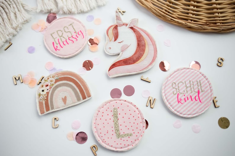 Peut inclure: Plusieurs &eacute;cussons en tissu aux couleurs pastel et des lettres en bois sur une surface blanche. Les &eacute;cussons repr&eacute;sentent un arc-en-ciel, une licorne et des motifs circulaires avec du texte et une lettre. Des confettis compl&egrave;tent la d&eacute;coration.