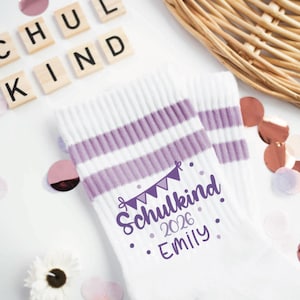 Könnte beinhalten: Weiße Sportsocken mit lila Streifen und dem Text "Schulkind 2026 Emily". Die Socken sind mit einem Banner-Grafik und Punkten verziert. Das Bild enthält Holzbuchstaben mit der Aufschrift "SCHUL KIND", Konfetti und einen Weidenkorb.