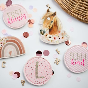 Könnte beinhalten: Eine Auswahl an dekorativen Aufnähern und Verzierungen, darunter ein Pferdekopf, ein Regenbogen und runde Aufnäher mit Text und Buchstaben. Die Aufnäher sind in Rosa-, Beige- und Brauntönen gehalten und mit floralen und glitzernden Akzenten versehen. Konfetti und Holzbuchstaben sind verstreut.