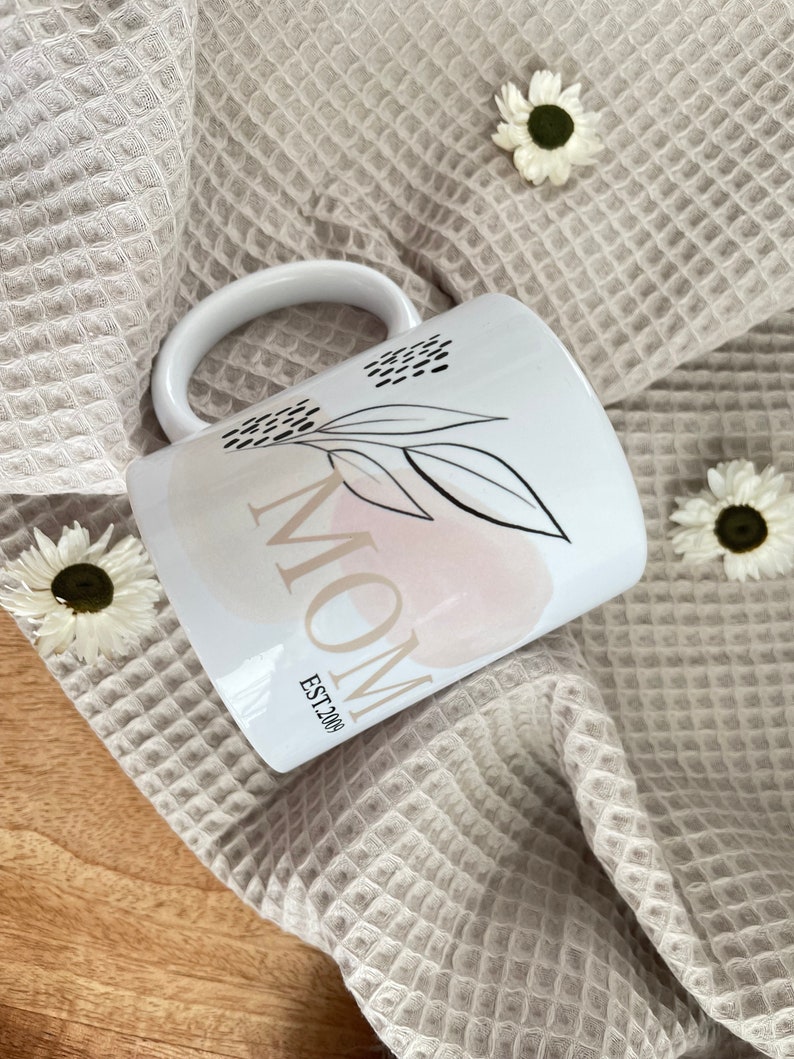 K&ouml;nnte beinhalten: Wei&szlig;e Keramiktasse mit einem abstrakten Design in Rosa und Wei&szlig; und dem Wort "MOM" mit "EST. 2004" darunter. Die Tasse steht auf einem beigefarbenen Waffelstoff.