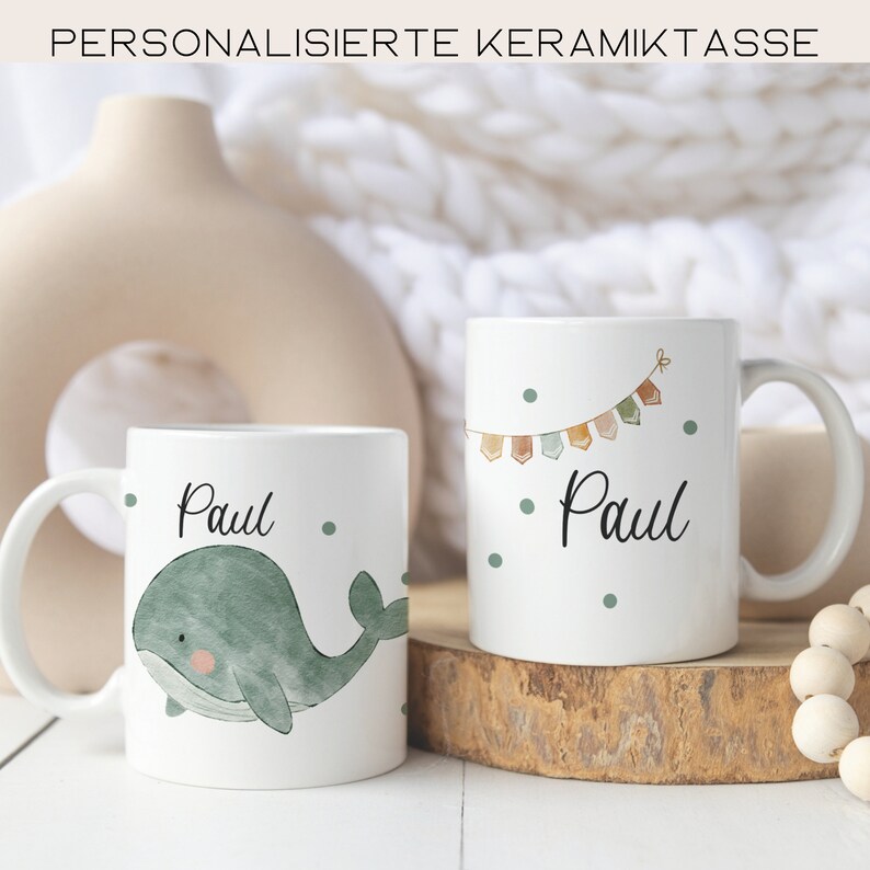 May include: Two white ceramic mugs with personalized names. One mug features a green whale illustration and the other has a string of colorful flags. Both mugs have green polka dots and the name "Paul" written in black.