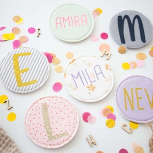 Könnte beinhalten: Sechs kreisförmige Stoffaufnäher mit personalisierten Namen und Buchstaben. Die Aufnäher umfassen ein mintgrünes mit rosa Text AMIRA, ein graues mit schwarzem M, ein grau gestreiftes mit goldenem E, ein florales mit lilafarbenem MILA, ein rosa gepunktetes mit goldenem L und ein lavendelfarbenes mit goldenem NEV.