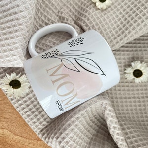K&ouml;nnte beinhalten: Wei&szlig;e Keramiktasse mit einem schwarz-wei&szlig;en Blumenmuster und dem Wort "MOM" in schwarzem Text mit den Worten "EST. 200" darunter.