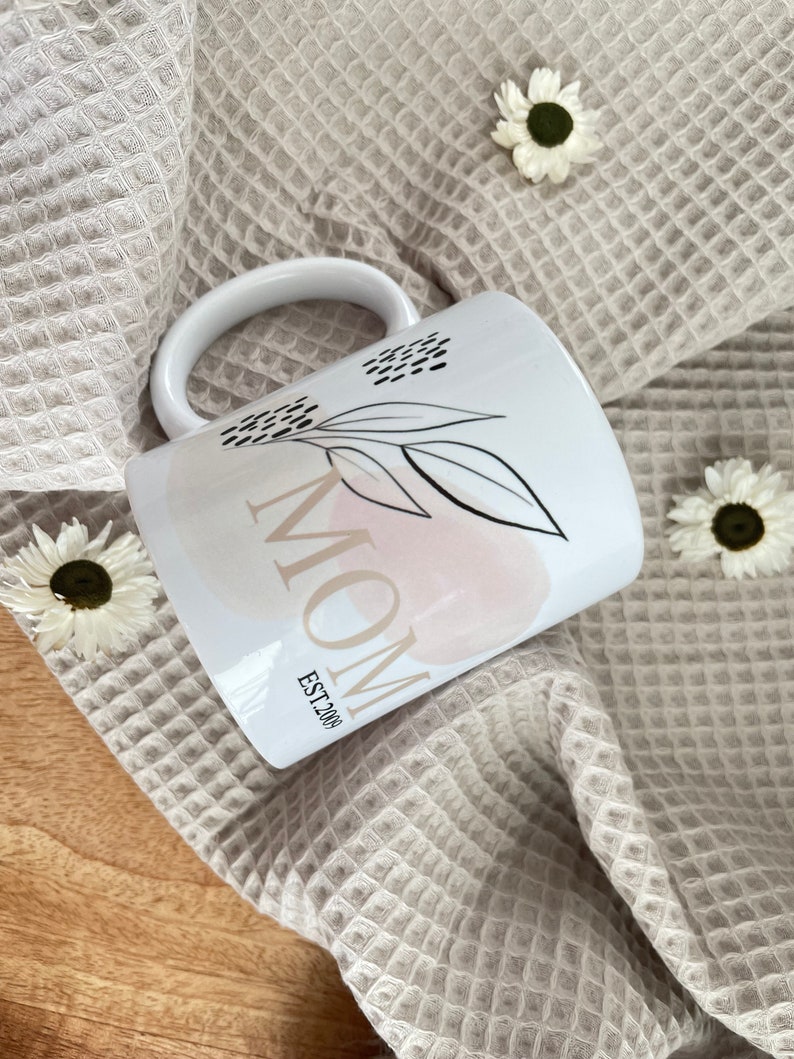K&ouml;nnte beinhalten: Wei&szlig;e Keramiktasse mit einem schwarz-wei&szlig;en Blumenmuster und dem Wort "MOM" in schwarzem Text mit den Worten "EST. 200" darunter.