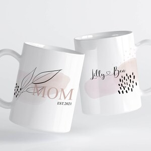 K&ouml;nnte beinhalten: Zwei wei&szlig;e Keramikbecher mit schwarzen und rosafarbenen Designs. Ein Becher sagt "MOM EST. 2021" und der andere sagt "Lilly Ben" mit einem Herzsymbol.