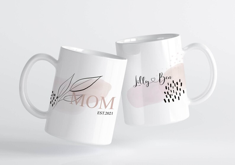 K&ouml;nnte beinhalten: Zwei wei&szlig;e Keramikbecher mit schwarzen und rosafarbenen Designs. Ein Becher sagt "MOM EST. 2021" und der andere sagt "Lilly Ben" mit einem Herzsymbol.