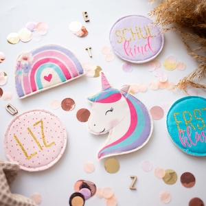 Könnte beinhalten: Eine Sammlung von Stoffaufnähern mit einem Regenbogen, einem Einhornkopf und runden Designs. Ein rosa Aufnäher zeigt den Namen LIZ in Goldglitzer, ein lila Aufnäher trägt die Aufschrift SCHUL kind und ein türkiser Aufnäher zeigt ERST klassig.