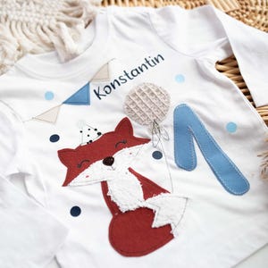 Könnte beinhalten: Weißes Langarmshirt mit einem Fuchsapplikation und dem Namen "Konstantin" in blauer Schrift. Das Shirt hat eine blaue "1" und dekorative Elemente, passend für einen Kindergeburtstag.