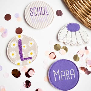Könnte beinhalten: Vier Stoffaufnäher, darunter ein runder beiger Aufnäher mit weißen Gänseblümchen und einem violetten Glitzerbuchstaben L, ein gestreifter beiger Aufnäher mit violettem Text, ein violetter runder Aufnäher mit silbernem Text MARA und eine Blütenform.
