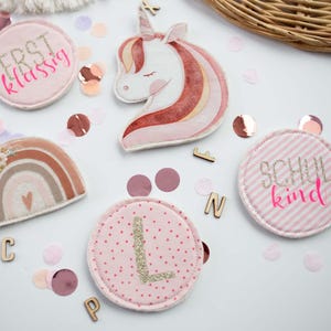 Peut inclure: Plusieurs &eacute;cussons en tissu rose avec des broderies. On y trouve une licorne, un arc-en-ciel et des &eacute;cussons circulaires avec du texte et une lettre. Des confettis et des lettres en bois sont &eacute;parpill&eacute;s.