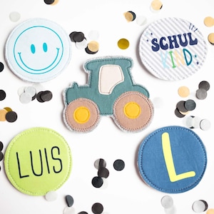 Könnte beinhalten: Ein Set aus fünf Stoffaufnähern, bestehend aus einem blaugrünen Traktor, einem hellblauen Kreis mit blaugrünem Smiley, einem weißen Kreis mit grauen Streifen und dem Text SCHULKIND, einem lindgrünen Kreis mit dem Namen LUIS und einem blauen Kreis mit dem Buchstaben L.