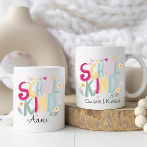 May include: Two white ceramic mugs with colorful text that reads "SCHOOL KIND 2026". One mug has the name "Anni" below the text. The other mug has the text "Du bist 1 Klasse".