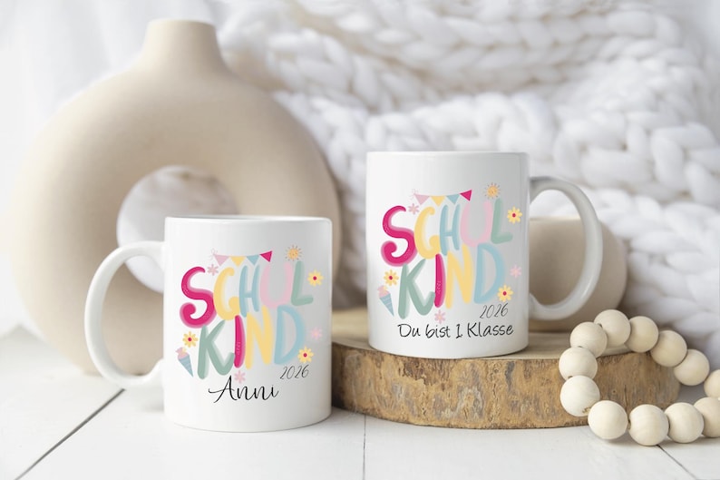 May include: Two white ceramic mugs with colorful text that reads "SCHOOL KIND 2026". One mug has the name "Anni" below the text. The other mug has the text "Du bist 1 Klasse".