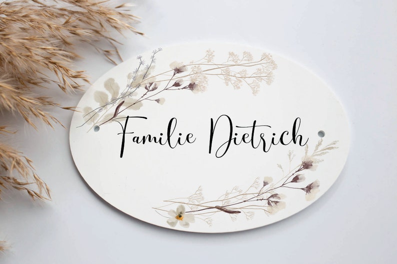 May include: White oval sign with a floral design and the text "Familie Dietrich" in black cursive lettering.