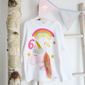 以下が含まれることがあります： 虹、ユニコーン、星柄の白い長袖シャツ。シャツには「Ella」と「6」がピンクとゴールドのキラキラで書かれています。