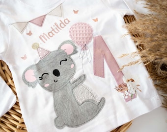 Könnte beinhalten: Ein weißes Langarmshirt mit einem rosa und grauen Koala, der einen rosa Ballon hält. Das Shirt ist mit dem Namen "Matilda" und einer rosa Eins bestickt.