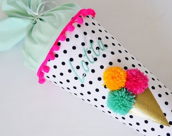 Op de afbeelding: Een witte en zwarte stippenkegel met een roze pomponrand. De kegel is geborduurd met de naam "Lotta" in groen garen. De kegel is versierd met drie pompons in geel, roze en groen.