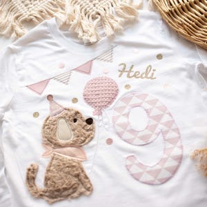 Könnte beinhalten: Weißes Langarmshirt mit Geburtstagsthema. Es zeigt einen Plüschhund mit Partyhut, einen rosa Ballon und die Zahl 9. Der Name "Hedi" ist in goldenem Glitzer über der Zahl geschrieben. Rosa und goldene Akzente vervollständigen das Design.