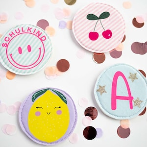 Könnte beinhalten: Vier bunte, runde Stoffaufnäher mit verschiedenen Designs. Einer zeigt ein Smiley-Gesicht mit dem Text "SCHULKIND", ein anderer Kirschen, ein dritter den Buchstaben "A" mit Sternen und der letzte eine Zitrone. Konfetti umgibt die Aufnäher.