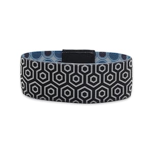 May include: A wristband featuring a black and white geometric hexagon pattern on the exterior. The interior lining displays a blue and dark blue circular pattern. A solid black rectangular tab secures the band at the top center.