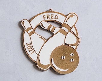 May include: A white wooden ornament with a bowling ball and two bowling pins. The ornament is decorated with the name "FRED" and the year "2021".