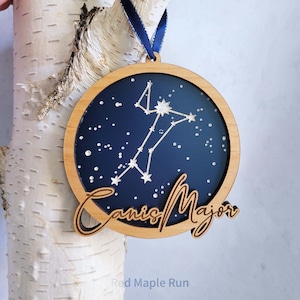 Puede incluir: Un adorno de madera con fondo azul y estrellas de purpurina plateada. La constelación Canis Major está representada en purpurina plateada. El adorno está etiquetado como "Canis Major" en una fuente cursiva.