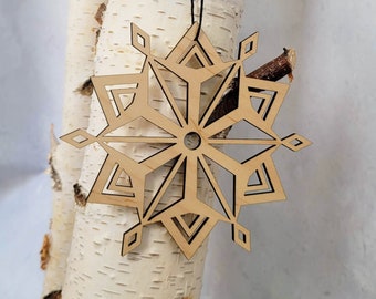 Op de afbeelding: Een houten sneeuwvlok ornament met een geometrisch ontwerp, hangend aan een berkenboomtak. Het ornament is lichtbruin van kleur en heeft een zwart touwtje.