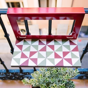 Op de afbeelding: Een rode metalen balkontafel met een wit, groen en rood geometrisch betegeld blad. De tafel is bevestigd aan een zwart metalen hek.