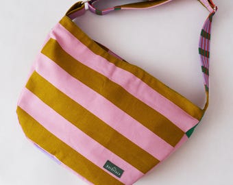 May include: A hobo-style shoulder bag featuring horizontal stripes in mustard yellow and light pink. The adjustable strap displays a pattern of lavender, green, and red stripes. A small rectangular green label with the text BULUPEACH is attached.