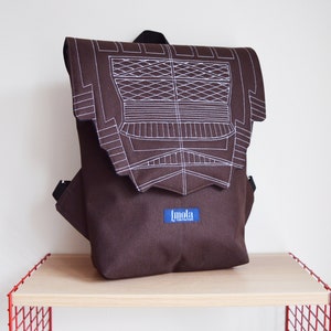 Puede incluir: Una mochila de lona marrón con un diseño geométrico blanco bordado en la solapa delantera. La mochila tiene una etiqueta azul con la palabra "Imola" en ella.