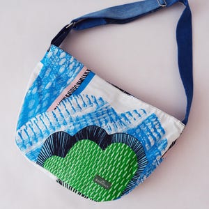 Op de afbeelding: Een schoudertas met een witte basis en een abstracte grafische print in blauw, groen en zwart. De tas heeft een lange, verstelbare donkerblauwe spijkerstof schouderband en een klein rechthoekig label met de tekst BOLDPEACH op de voorzijde.