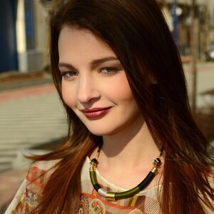 以下が含まれることがあります： 長い茶色の髪の女性が、カラフルなペイズリー柄のシャツに黒と緑のビーズネックレスを着用している。