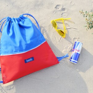 May include: A red and blue drawstring bag with a zippered pocket and the word "Imala" on the front. The bag is on a sandy beach with a pair of yellow sunglasses and a can of Red Bull.