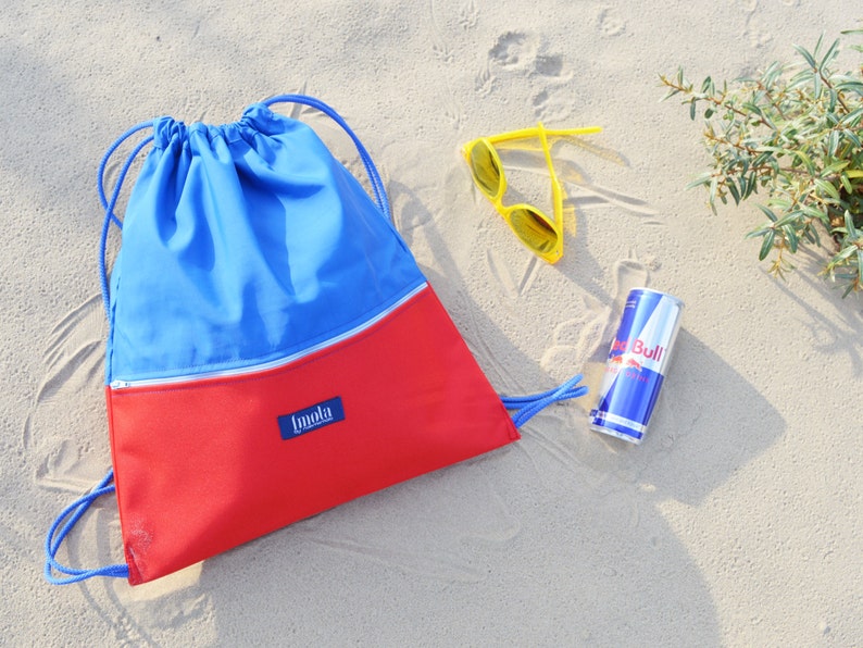 May include: A red and blue drawstring bag with a zippered pocket and the word "Imala" on the front. The bag is on a sandy beach with a pair of yellow sunglasses and a can of Red Bull.