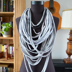 Può includere: Una sciarpa infinita grigia e bianca realizzata con t-shirt riciclate. La sciarpa è a strati e drappeggiata su un manichino nero.