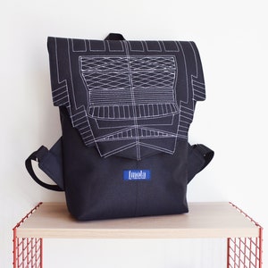 Puede incluir: Mochila de lona negra con un diseño geométrico blanco bordado en la solapa delantera. La mochila tiene una etiqueta azul con la palabra "Imola" en ella.