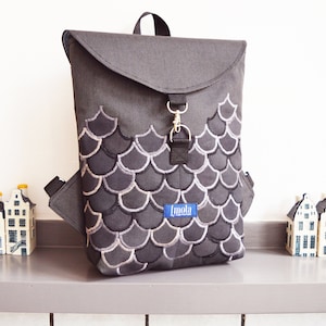 Peut inclure: Un sac à dos gris avec une fermeture à rabat et un motif d'écailles noir et blanc. Le sac à dos a une petite poche sur le côté et une étiquette bleue qui dit "Imola".