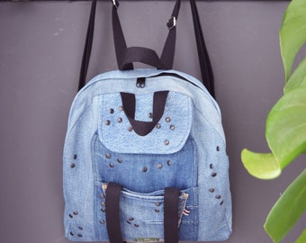 May include: A denim backpack with black straps and a front pocket. The backpack is adorned with dark metal studs and features a small pocket with a label. The bag is hanging on a black wall with silver hooks.