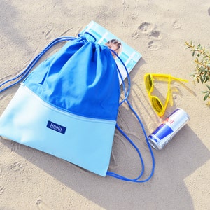 May include: A blue and light blue drawstring bag with a white zipper and the word "Imala" on the front. The bag is lying on a sandy beach with a pair of yellow sunglasses and a red and blue can.