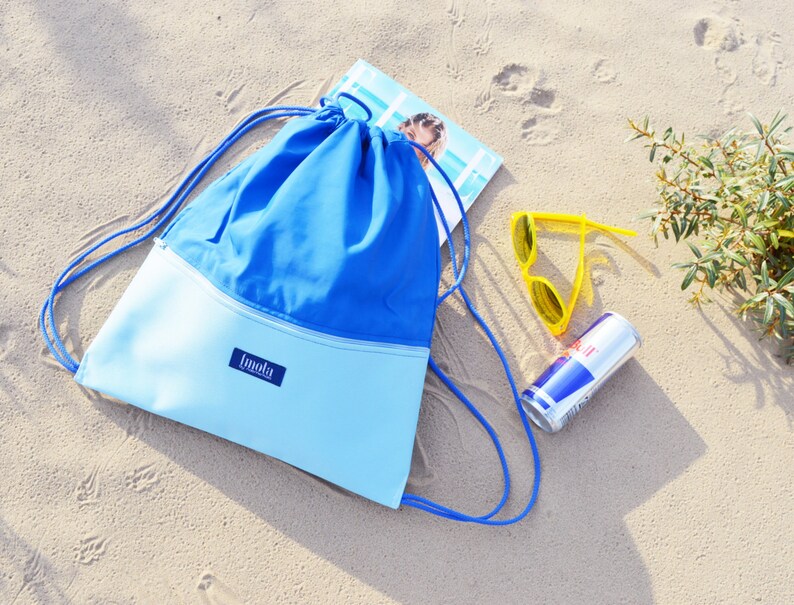 May include: A blue and light blue drawstring bag with a white zipper and the word "Imala" on the front. The bag is lying on a sandy beach with a pair of yellow sunglasses and a red and blue can.