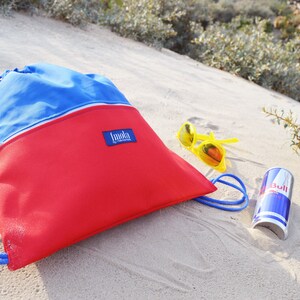 May include: A red and blue drawstring bag with the word "Immoka" on the front, yellow sunglasses, and a red can of Red Bull on a sandy beach.