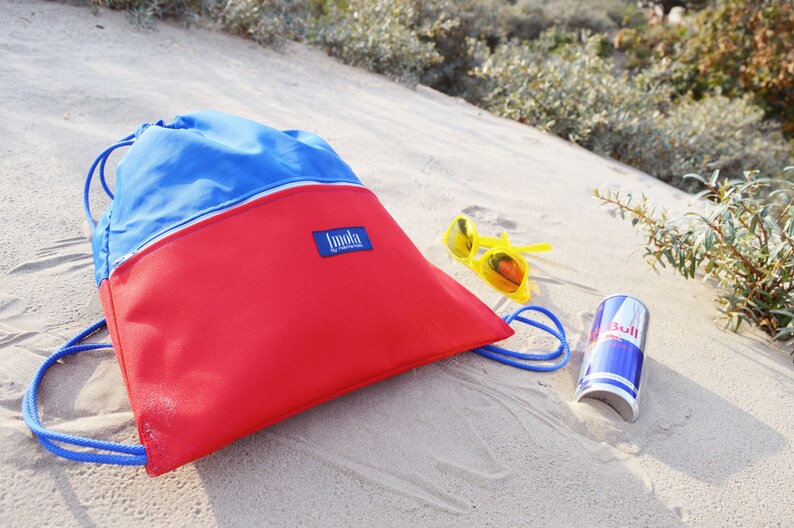 May include: A red and blue drawstring bag with the word "Immoka" on the front, yellow sunglasses, and a red can of Red Bull on a sandy beach.