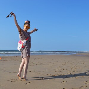 K&ouml;nnte beinhalten: Eine Person am Sandstrand mit erhobenen Armen, die eine Sonnenbrille h&auml;lt. Sie tr&auml;gt ein taupefarbenes Kleid und eine wei&szlig;e Tasche mit floralem Design. Der Himmel ist strahlend blau, und das Meer ist im Hintergrund zu sehen.