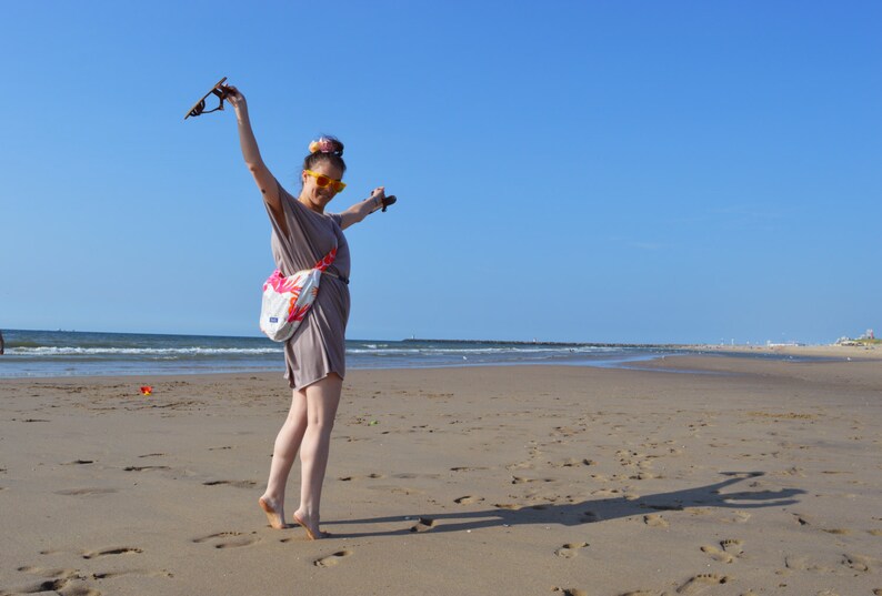 K&ouml;nnte beinhalten: Eine Person am Sandstrand mit erhobenen Armen, die eine Sonnenbrille h&auml;lt. Sie tr&auml;gt ein taupefarbenes Kleid und eine wei&szlig;e Tasche mit floralem Design. Der Himmel ist strahlend blau, und das Meer ist im Hintergrund zu sehen.