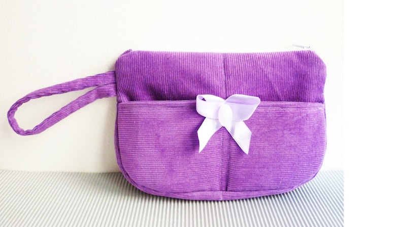 May include: A small purple corduroy purse with a white bow and a handle.