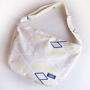 Könnte beinhalten: Beige Umhängetasche mit einem geometrischen Muster aus gelben, weißen, rosa und blauen Linien und Formen. Die Tasche hat einen passenden verstellbaren Riemen und den Markennamen "Imola" auf einem blauen Etikett.