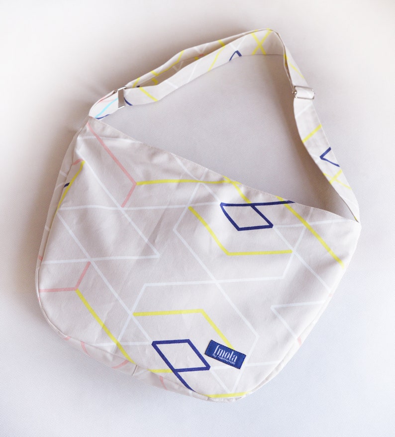 K&ouml;nnte beinhalten: Beige Umh&auml;ngetasche mit einem geometrischen Muster aus gelben, wei&szlig;en, rosa und blauen Linien und Formen. Die Tasche hat einen passenden verstellbaren Riemen und den Markennamen "Imola" auf einem blauen Etikett.