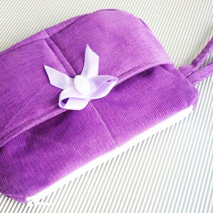 May include: A purple corduroy zippered pouch with a white zipper and a purple satin bow. The pouch has a handle and is lying on a white striped surface.