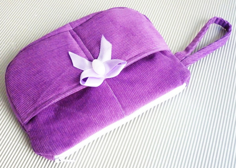 May include: A purple corduroy zippered pouch with a white zipper and a purple satin bow. The pouch has a handle and is lying on a white striped surface.